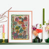 Lucy Innes Williams painting of dark pink and orange flowers in a pink frame it is propped on a green shelf next to two candlesticks with a green and a pink candle 