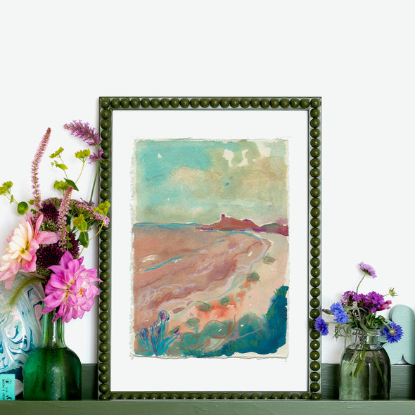 Framed Lucy Innes Williams painting in pinks and greens of beach and headland it is propped on shelf next to green glass bottles filled with pink flowers