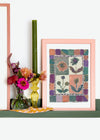 Framed  Lucy Williams  painting of flowers enclosed by geometric square in pink and green, the painting is propped on a green shelf with a yellow glass candlestick and green candle 