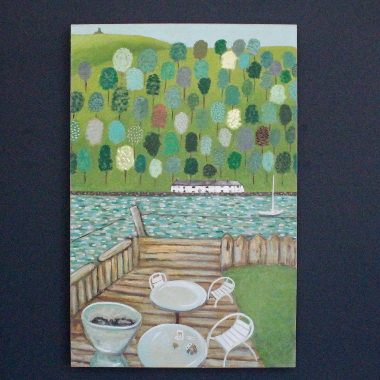 White chairs and tables set on the marina looking out to sea, with multicoloured trees and white houses on the peninsula by UK artist Siobhan Purdy.