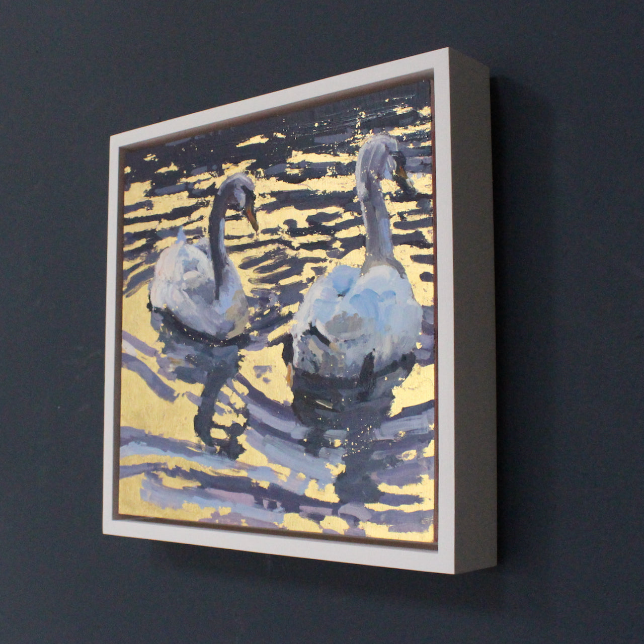 A  framed painting of two swans swimming on gold water by Jill Hudson