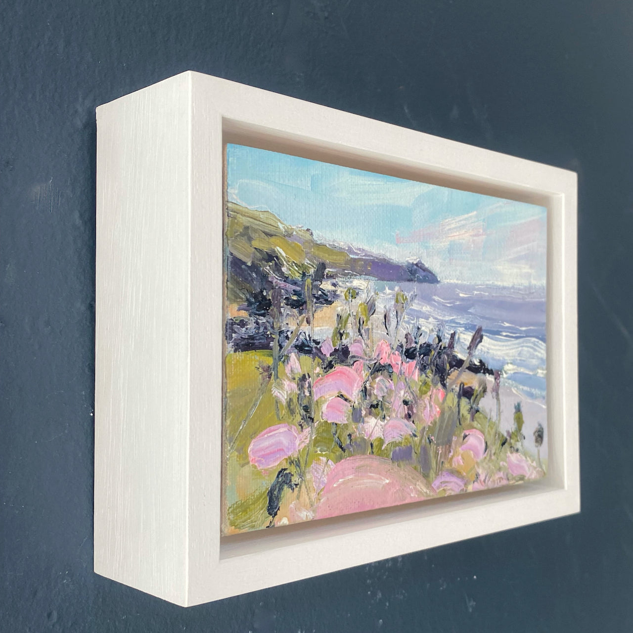 A painting of Rame Head in Cornwall by Jill Hudson, the headland is dark green under a pale blue sky, the sea is purple and in the foreground there are pale pink wildflowers. The painting is in a white  frame 