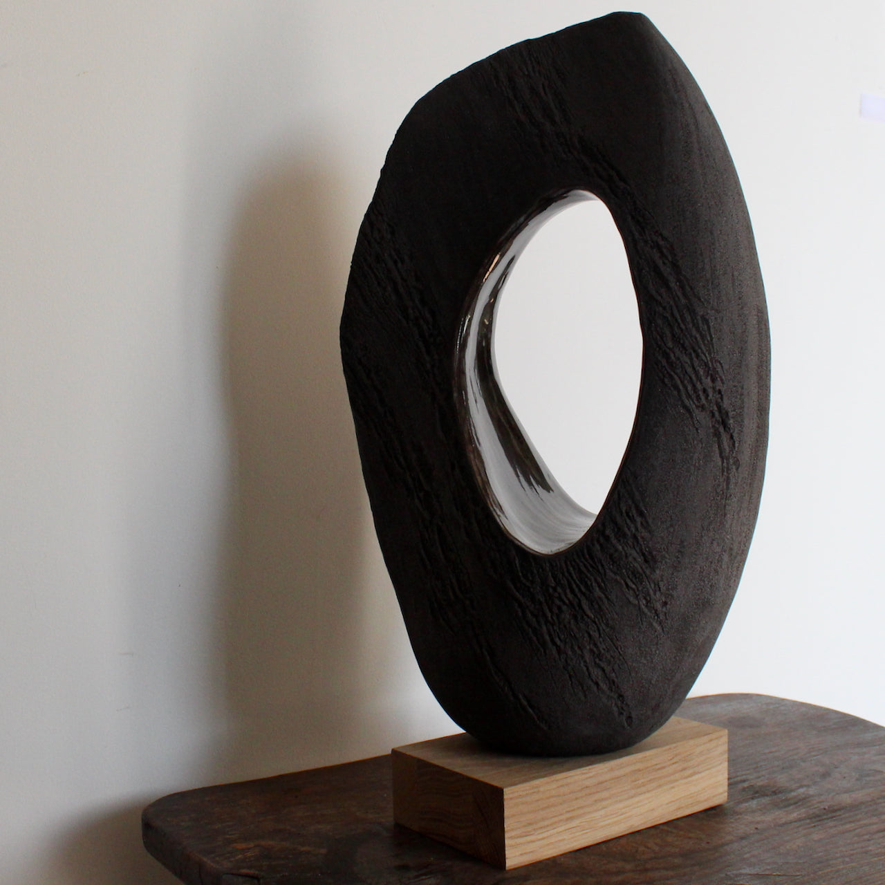 An Anthea Bowen brown oval shaped sculpture with hollow centre on an oak stand on top of a table.