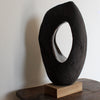 An Anthea Bowen brown oval shaped sculpture with hollow centre on an oak stand on top of a table.