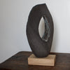Oval brown sculpture with hollow centre on an oak stand by Cornish artist, Anthea Bowen.