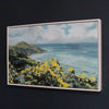 a framed painting by Cornish artist Imogen Bone of Rame Head in Cornwall with yellow & green foliage with green coastline and blue sea to the left, blue sky with white clouds 