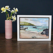 a small framed Jill Hudson painting of Rame Head in Cornwall with a golden beach, dark rocks and green headland in the blue sea.