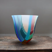 a tall and gently curved multi-coloured glass vase on a wooden table it is made by glass artist Ruth Shelley.