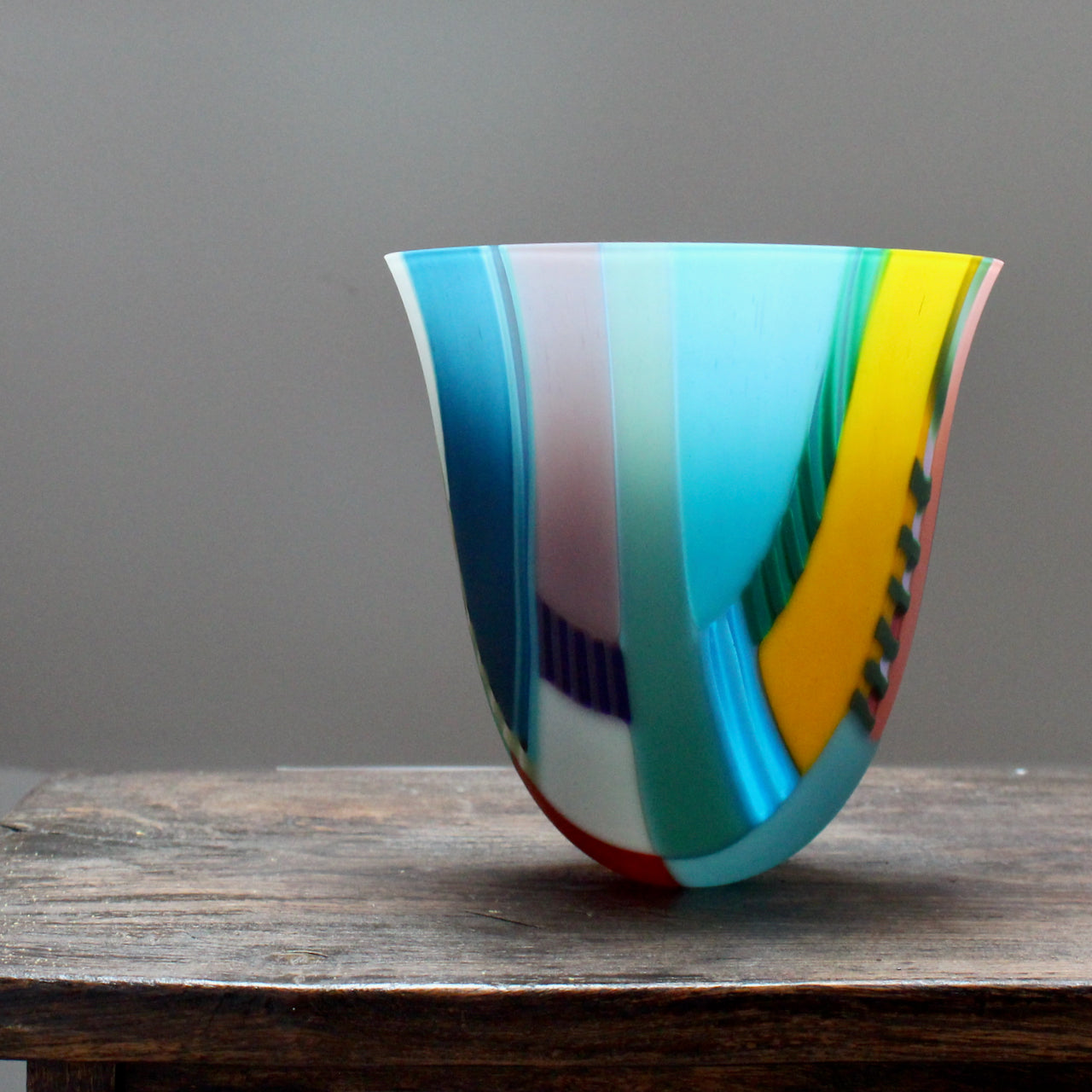 a tall and gently curved multi-coloured glass vase on a wooden table by Ruth Shelley