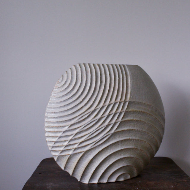 Michele Bianco pale white stoneware ceramic disc shaped vessel with carved lines by pictured on a dark wood table