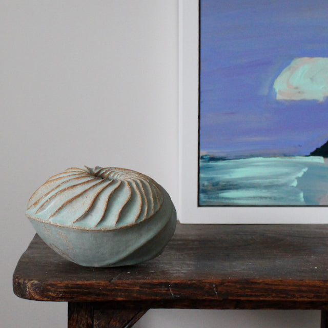 A round and carved pale green ceramic vessel by Michele Bianco photographed on a dark wood table with the corner of a purple and green painting 