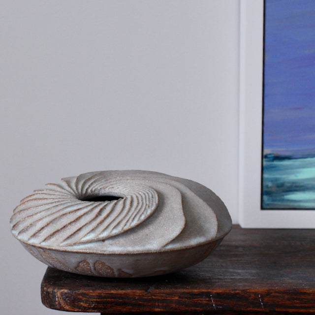 A round and carved ceramic vessel by Michele Bianco photographed on a dark wood table with the corner of a purple and green painting just visible. 