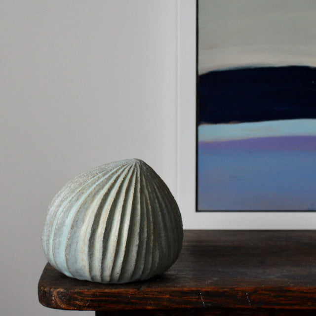 pale green carved stoneware ceramic pod shaped sculpture by Michele Bianco on a dark wooden table with the corner  of a painting visible