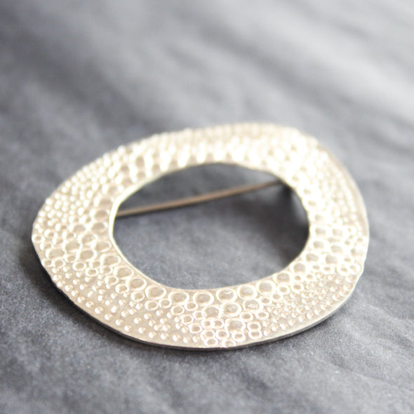 a textured silver round brooch with a hollow middle by UK jewellery designer Ann Bruford