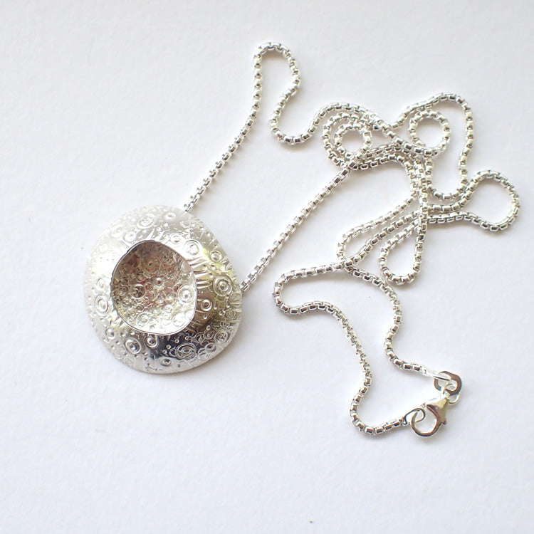 large silver pendant with a delicate circle design on the silver and an indent in the centre it's on a silver chain  and is by jewellery designer Ann Bruford