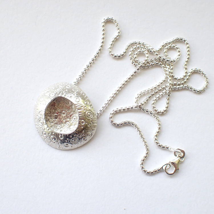 large silver pendant with a delicate circle design on the silver and an indent in the centre it's on a silver chain  and is by jewellery designer Ann Bruford 