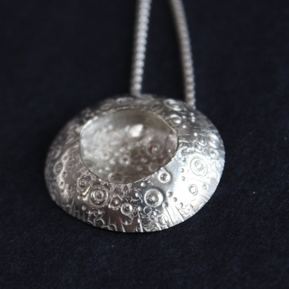 a large silver pendant with a delicate circle design on the silver and an indent in the centre it's on a silver chain  and is by jewellery designer Ann Bruford.