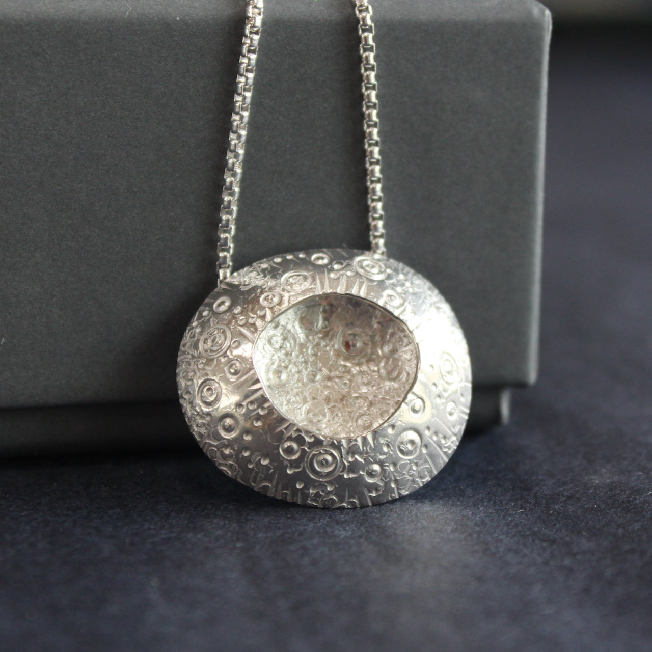 large silver pendant with a delicate circle design on the silver and an indent in the centre it's on a silver chain  and is by jeweller Ann Bruford.