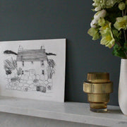 a black and white print by Sophie Harding  of a Cornish Cottage it is resting on a marble mantle piece next to a glass vase and  some flowers 