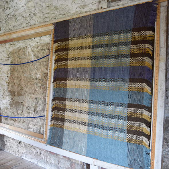 woollen throw by Rhain Wyman Design in shades of purple, blue and yellow shown on a loom 
