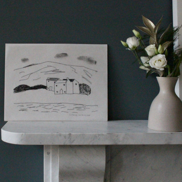 a black and white Sophie Harding print of cottages behind the sea and infront of a hill it is places on a mantlepiece next to a vase with flowers in it.