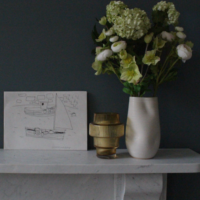 a drawing by Sophie Harding of a sailing boat in a harbour it is resting on marble top next to a glass vase and a taller white vase with  flowers in it