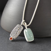 a double silver pendant on a silver chain with one green stone and one small orange topaz it is by  Carin Lindberg. 