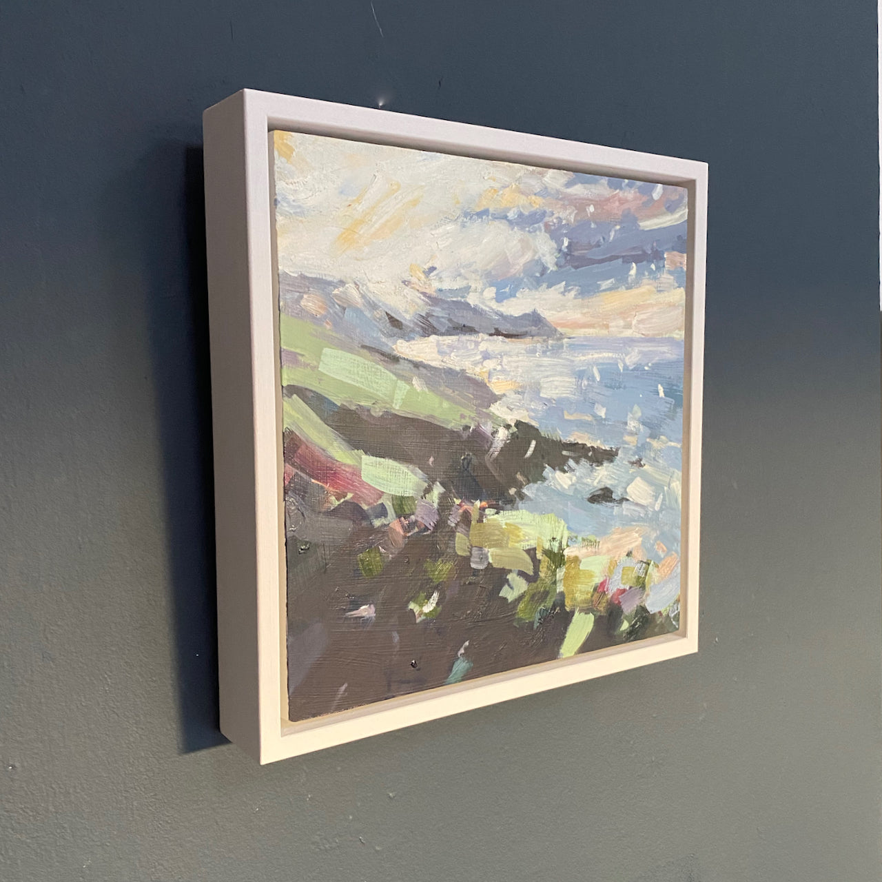 a framed Jill Hudson painting of Rame Head coastline in Cornwall in a wintery scene with a flurry of snowflakes the painting is hanging on a dark blue wall