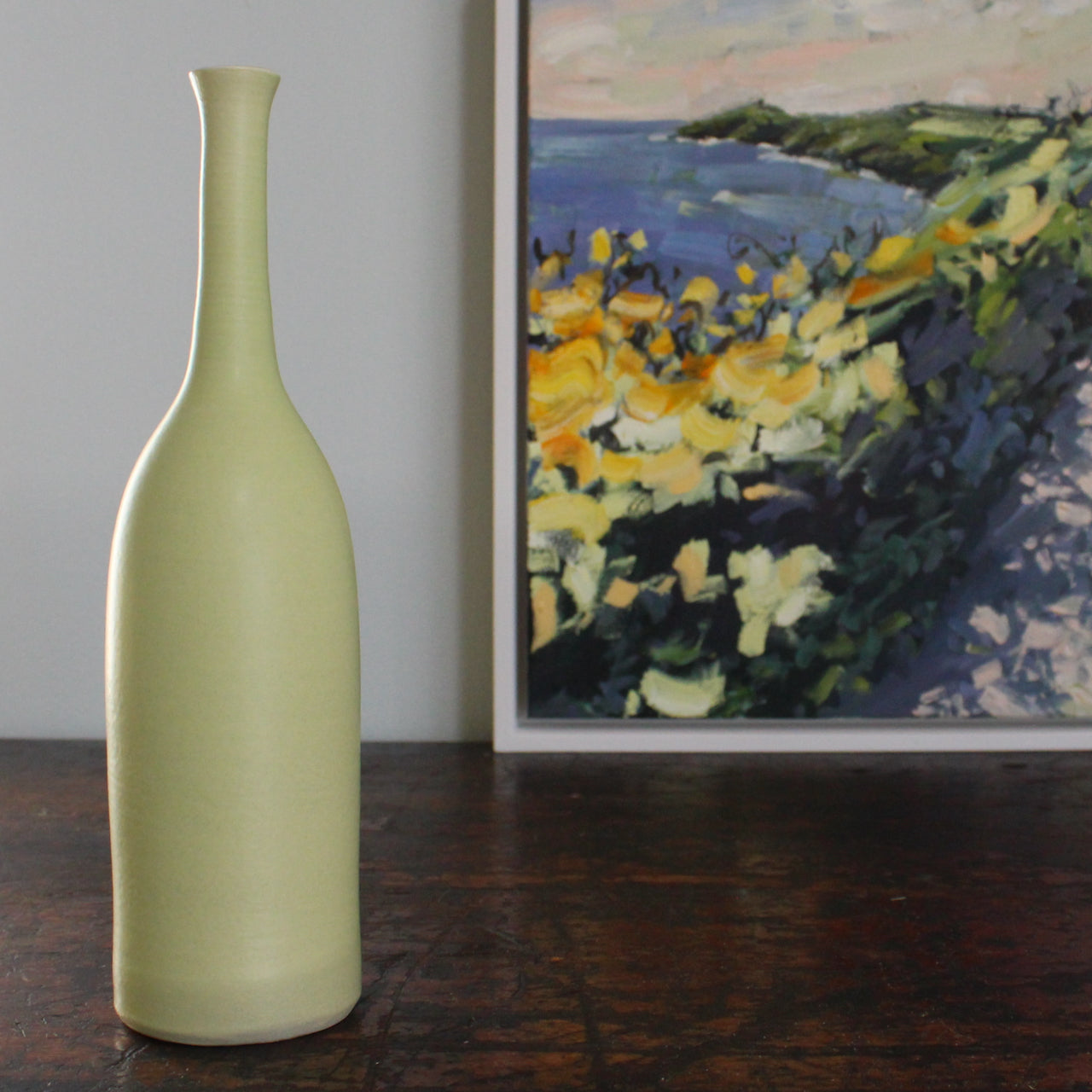 a ceramic bottle in  almond green by UK potter Lucy Burley next to the corner of a painting of Rame Head in Cornwall by artist Jill Hudson