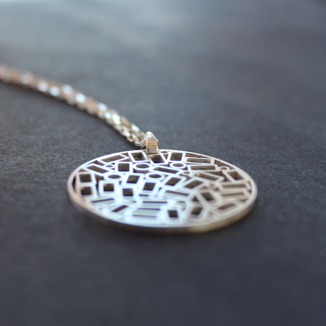 A round silver pendant on a chain, the centre is made up of cut out rectangles and small circles