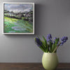 Oil painting of  a field by Jill Hudson, hanging on a grey wall. Green vase with purple flowers by Lucy Burley under it.