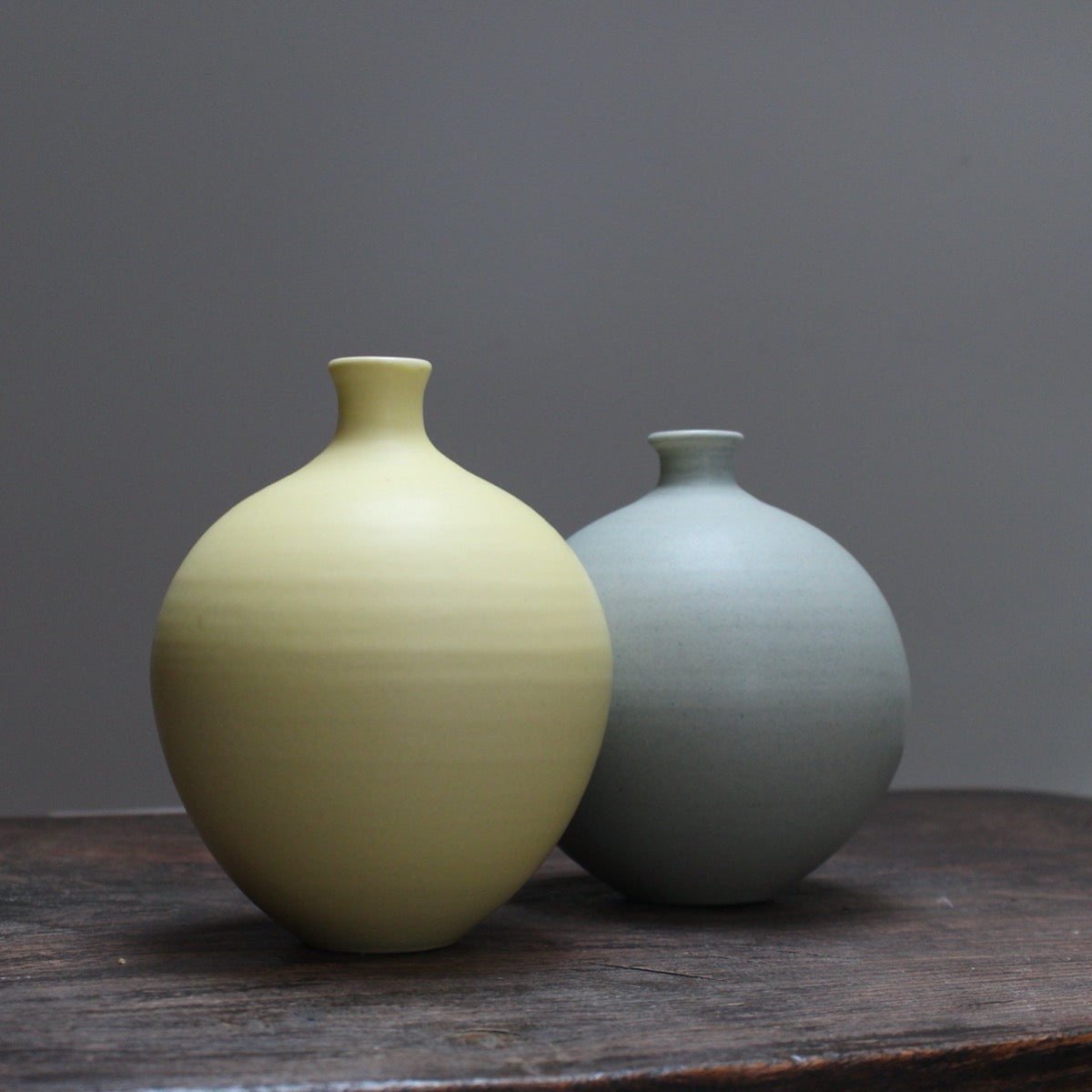 Two round ceramic bottles, one in pale yellow and one light sage on a wooden table at the Byre Gallery
