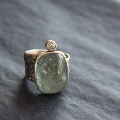 a silver ring with an oval shaped pale blue aquamarine stone and smaller diamond on a textured silver band by Cornwall jeweller Carin Lindberg