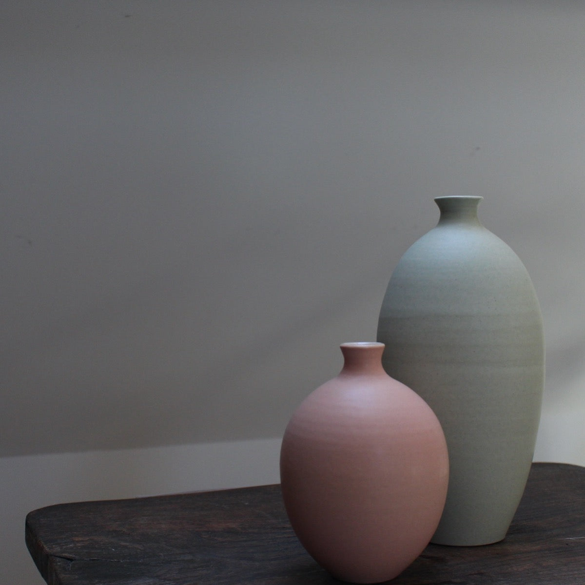 Two ceramic bottles by Lucy Burley on a wooden table - an oval shaped pale pink one and a taller one in light green shade