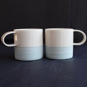 a pair of small espresso style cups by Kathryn Sherriff of By the Line Pottery they are in white porcelain with a turquoise ring design around the bottom half.