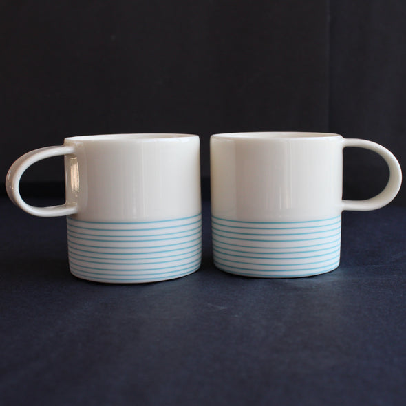 a pair of small espresso style cups by Kathryn Sherriff of By the Line Pottery they are in white porcelain with a turquoise ring design around the bottom half.