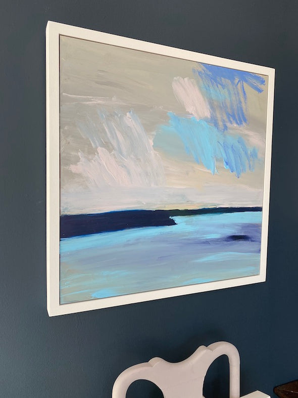 framed abstract painting of dark rocks pale blue and purple beach and sky by Cornwall artist Alex Yarlett it is hanging on a dark wall above a grey chair 