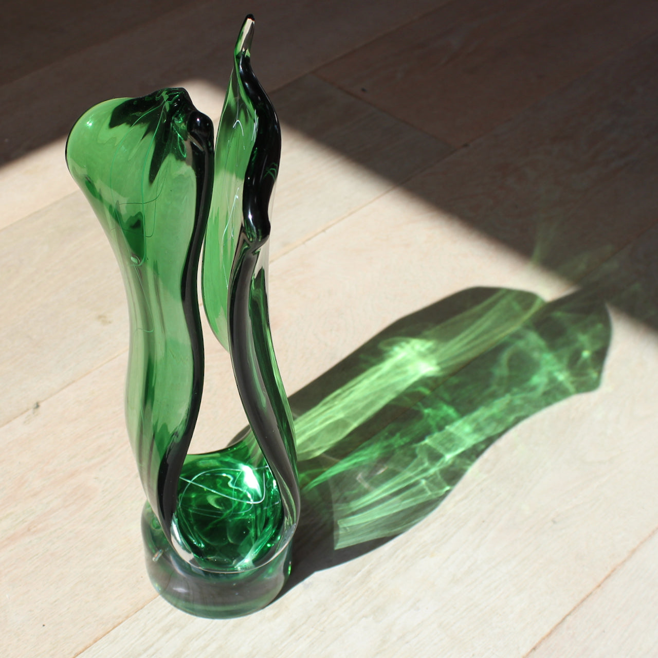 Benjamin Lintell glass sculpture in green photographed in the sunlight so that it casts a green shadow on to a pale wooden floor