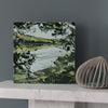 Imogen Bone painting of a pale blue river framed by a tree and the green fields on the far side the painting is on a marble mantlepiece infront of a grey wall
