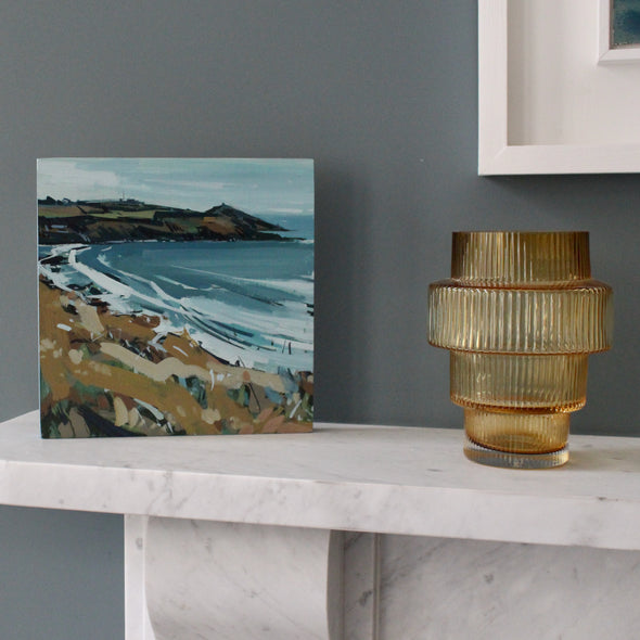 Imogen Bone landscape painting of Rame Head, a peninsula in south east Cornwall, painted in autumn with a blue sea, pale sky and brown and green grasses , the painting is standing on marble fireplace next to a gold coloured glass vase 
