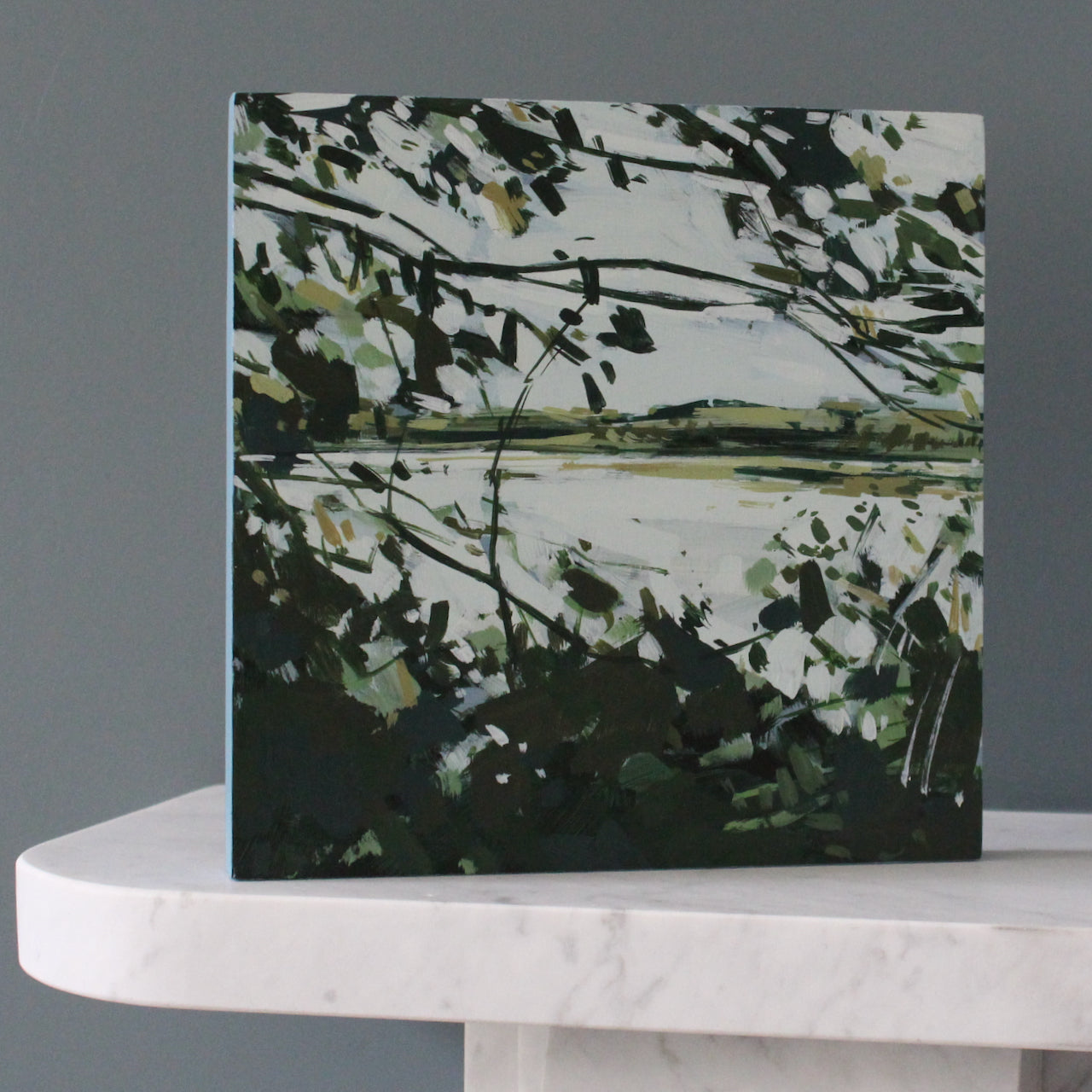 Imogen Bone painting looking through leaves and branches of a tree to a pale river with soft green fields on the other side, the painting is standing on a marble mantle piece in front of a grey wall.