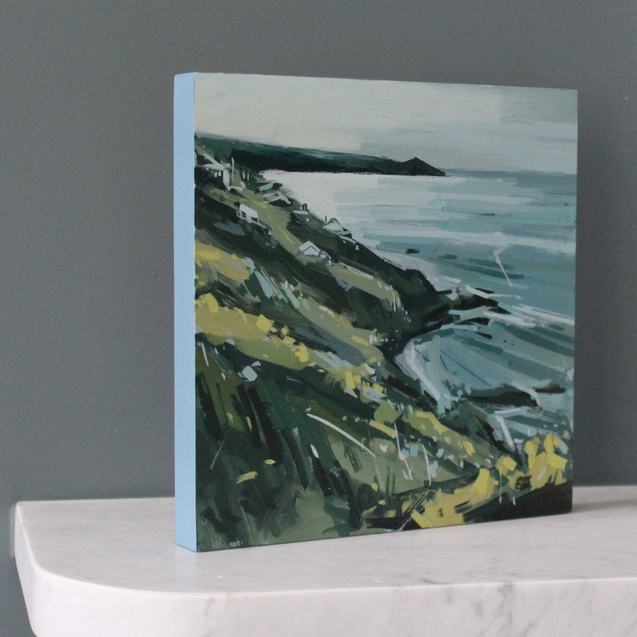 a Painting by Imogen Bone, Cornwall artist of the village of Freathy on the coast in south Cornwall looking towards Rame Head; the sky and sea are a soft wintery blue and the land dark green with bursts of yellow -  the painting is sitting on a marble mantle shelf in front of a grey wall