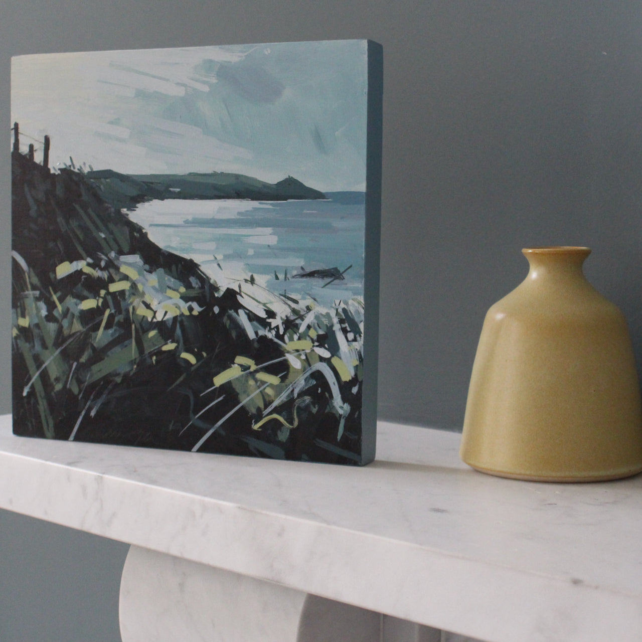 Imogen Bone landscape painting of Rame Head, a peninsula in south east Cornwall, the headland is dark green as are the grasses in the foreground of the painting, the painting is sitting on a marble fireplace next to a small yellow bottle