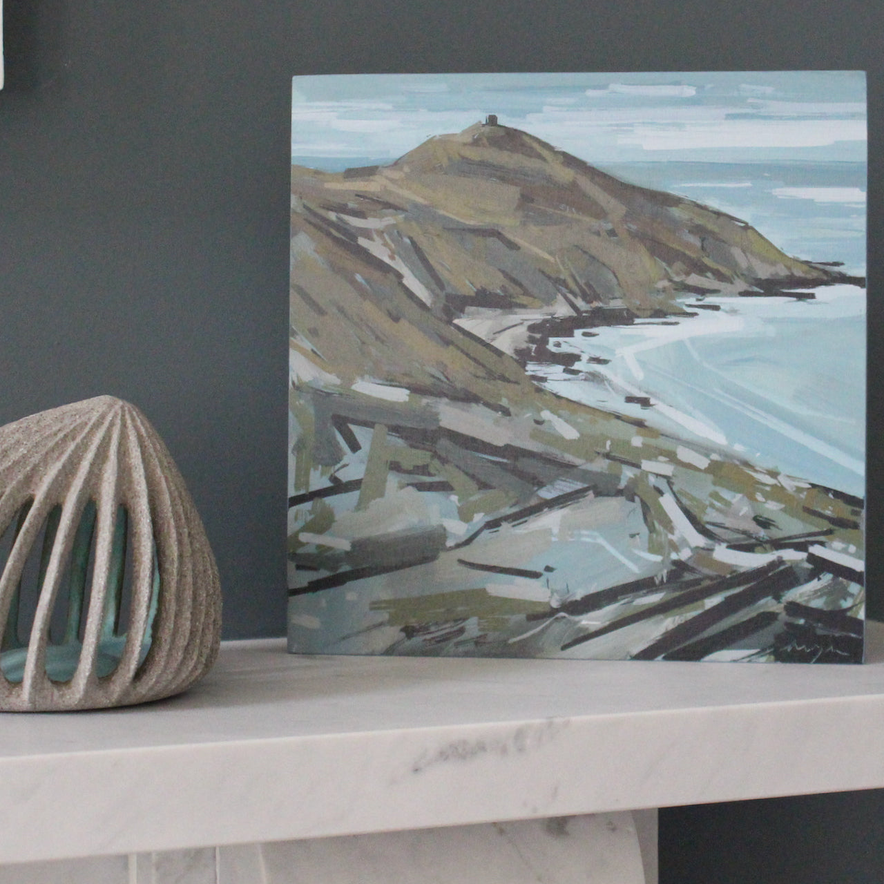 Painting of Rame Head in south east Cornwall in autumn colours with a pale blue sea and sky painted by Imogen Bone  - the painting is standing on a marble mantle piece next to small ceramic sculpture 