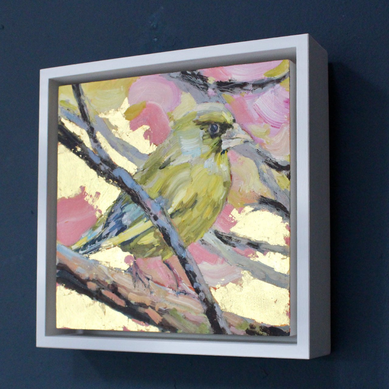 a framed Jill Hudson painting of a greenfinch on a branch with a pink, yellow and gold background.