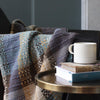 a woollen throw by Rhain Wyman Design in shades of purple, blue and yellow shown over the arm of a chair next to table with a mug on top of two books