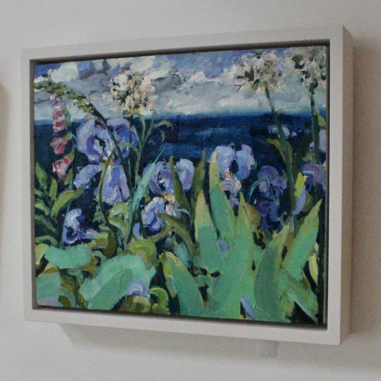 a framed Jill Hudson oil painting of purple iris flowers and pink lupins in a garden overlooking a blue sea.