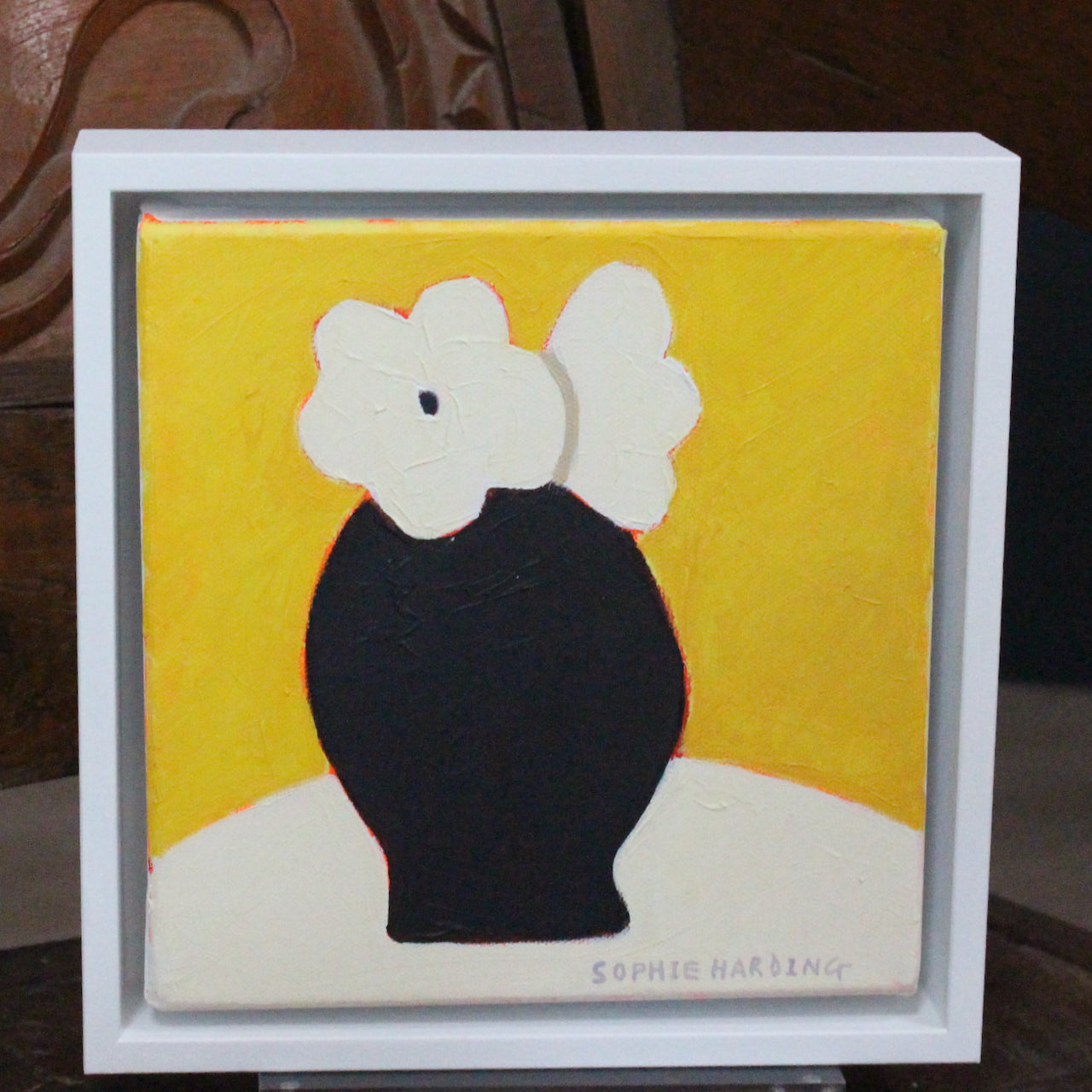 A framed Sophie Harding painting of white flowers in a black vase against a yellow background.
