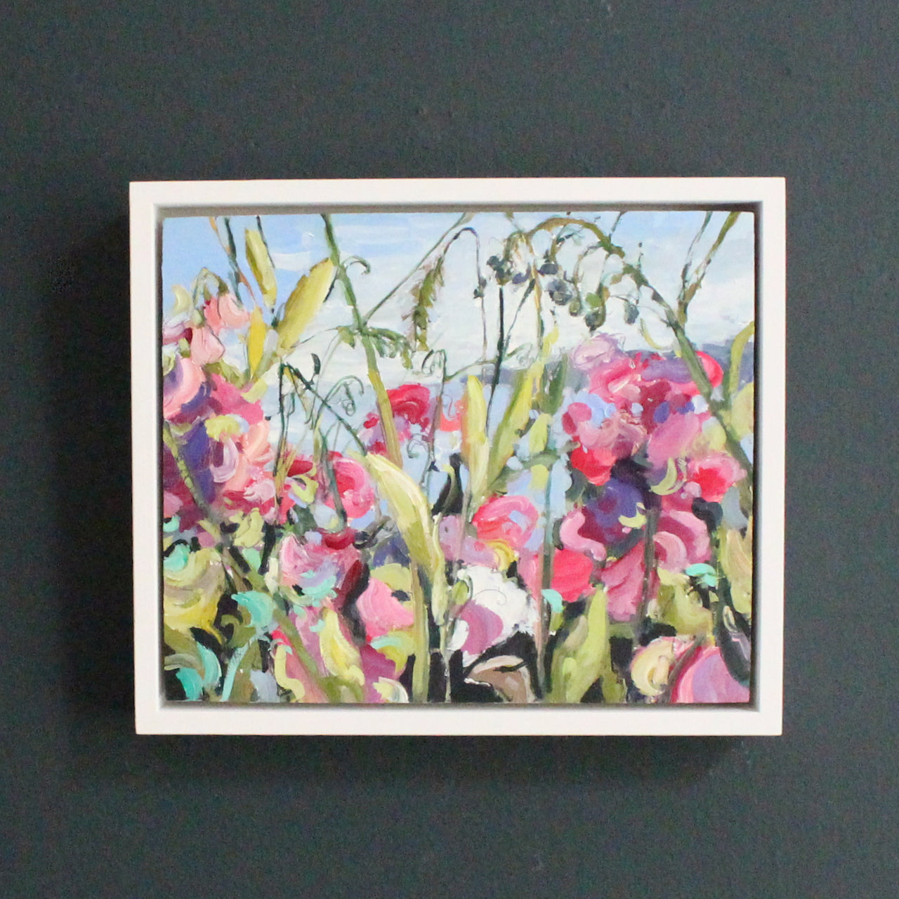 a Jill Hudson oil painting of  pink flowers and grasses with a glimpse of the sea and coastline visible , it is framed with a white frame