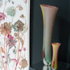 an extra tall glass vase by Ruth Shelley, UK glass artist in shades of pink, yellow and green it is next to a smaller vase in similar colours and both are positioned by a large floral painting 